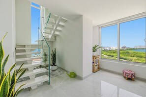 A modern interior with a glass staircase, a large window with a view, and a small plant on the floor. - A360.W62 -Atlantic Haven 1BR Penthouse (Noord)