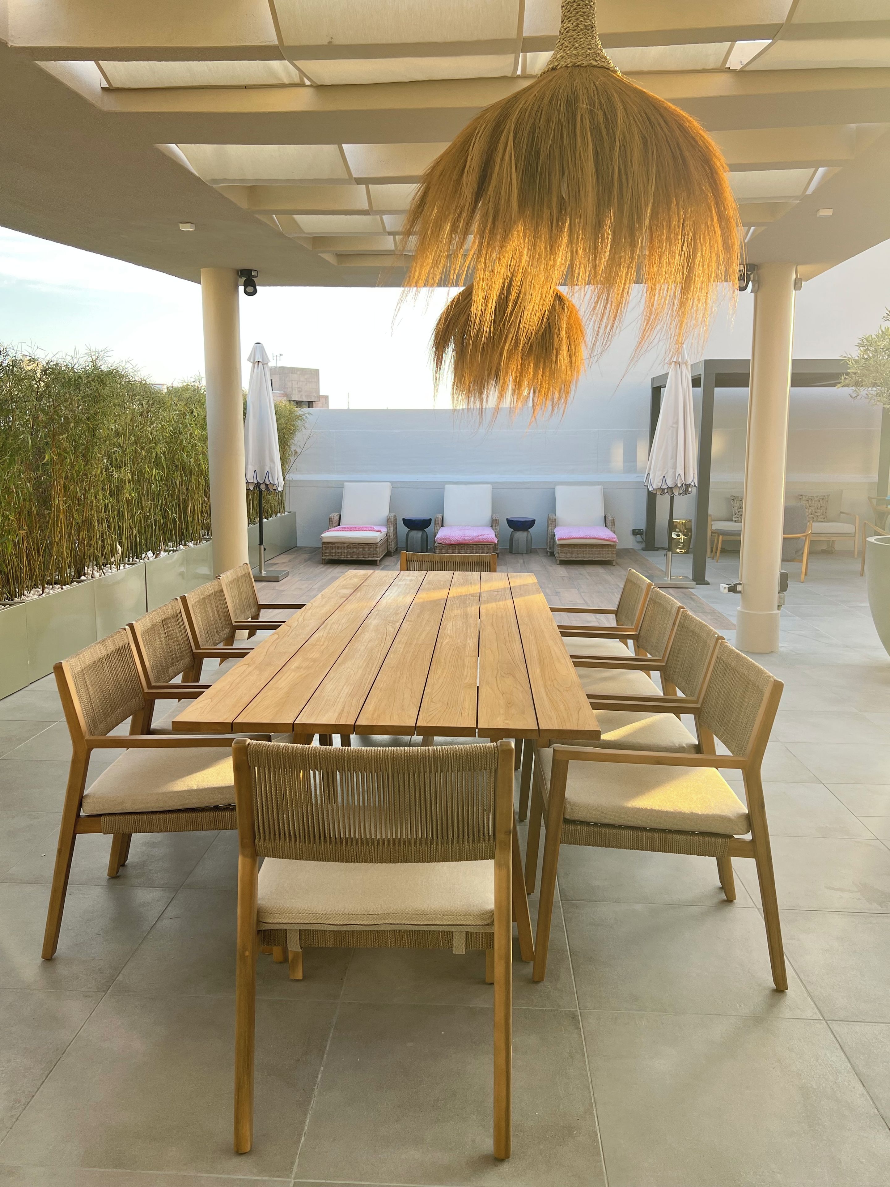 Un patio al aire libre con mesa y sillas de madera, una lámpara colgante y una zona de estar con cojines.