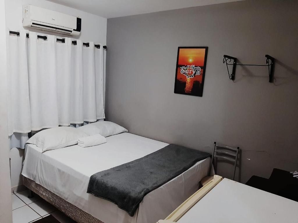 Un dormitorio con cama, cortinas blancas, un equipo de aire acondicionado, un cuadro en la pared y una mesita.