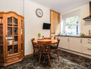 A kitchen with a wooden dining table, chairs, a cabinet with glass doors, a wall-mounted clock, and a window with a view. - Derwen-las (Penmaenmawr)