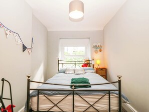 A bedroom with a bed, a window, a bedside lamp, and a wall-mounted shelf. - Derwen-las (Penmaenmawr)