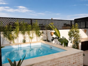 A swimming pool with a clear blue water surface, surrounded by a brick wall and various plants. - Riad privé MASKAT (Marrakech)