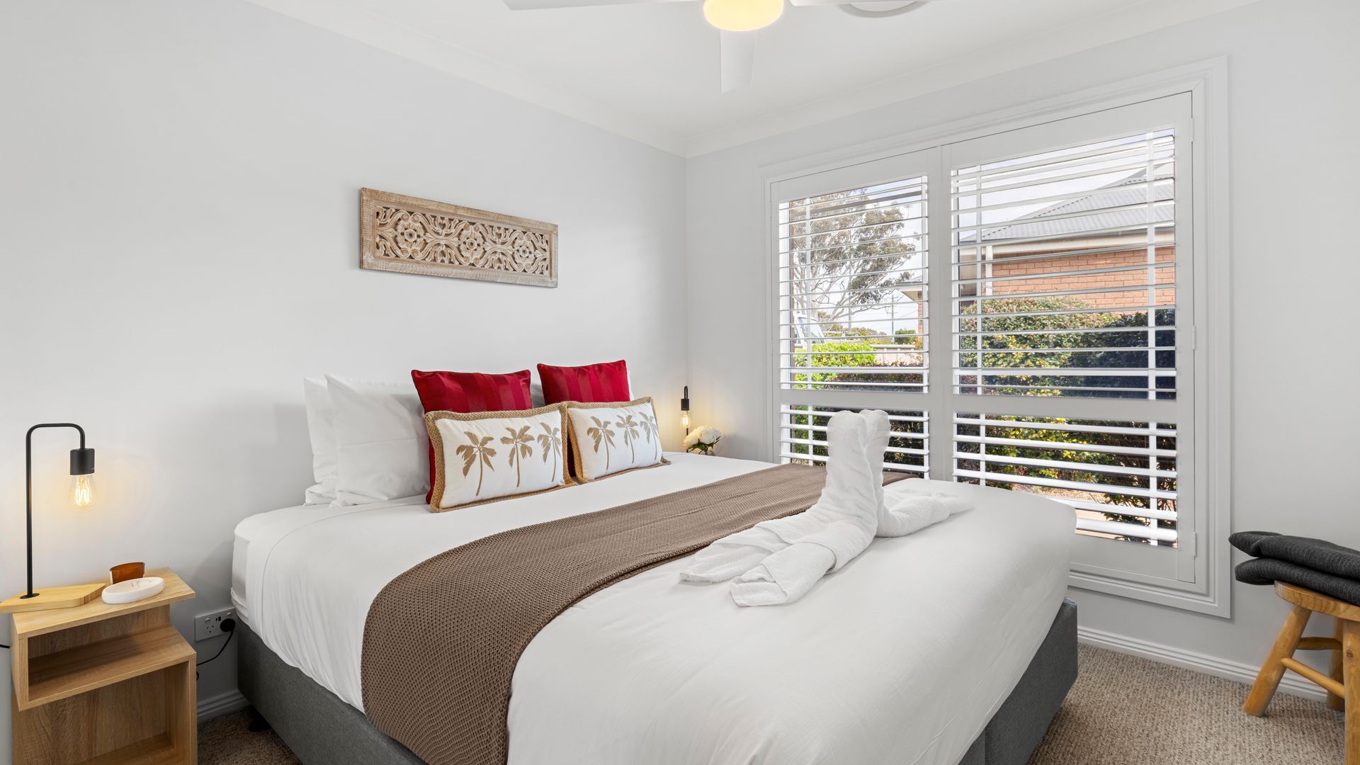 A hotel room with a bed, a bedside table with a lamp, a window with blinds, and a view of greenery outside.
