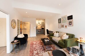 Un salon moderne avec un canapé vert, un fauteuil noir et une table basse. On aperçoit une cuisine au fond.
