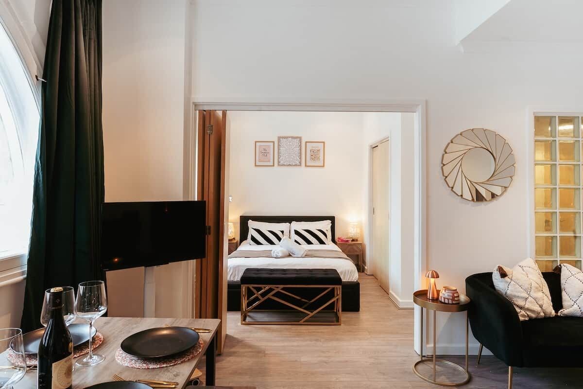 Ein Hotelzimmer mit einem Bett, einem Esstisch mit Weingläsern, einem Fernseher und einem schwarzen Sofa.