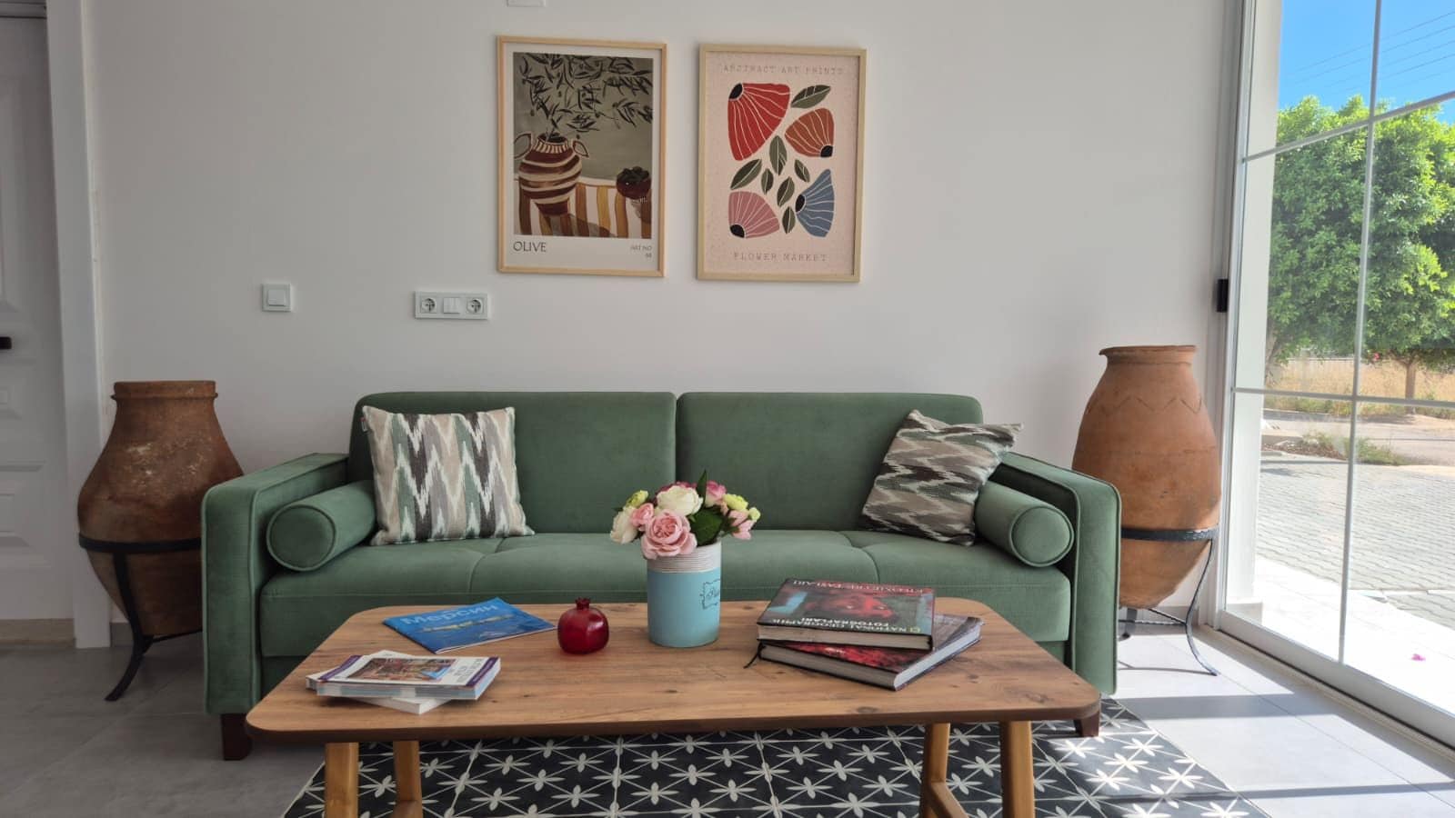 A living room with a green sofa, a coffee table with books and a vase of flowers, and two wall-mounted artworks.