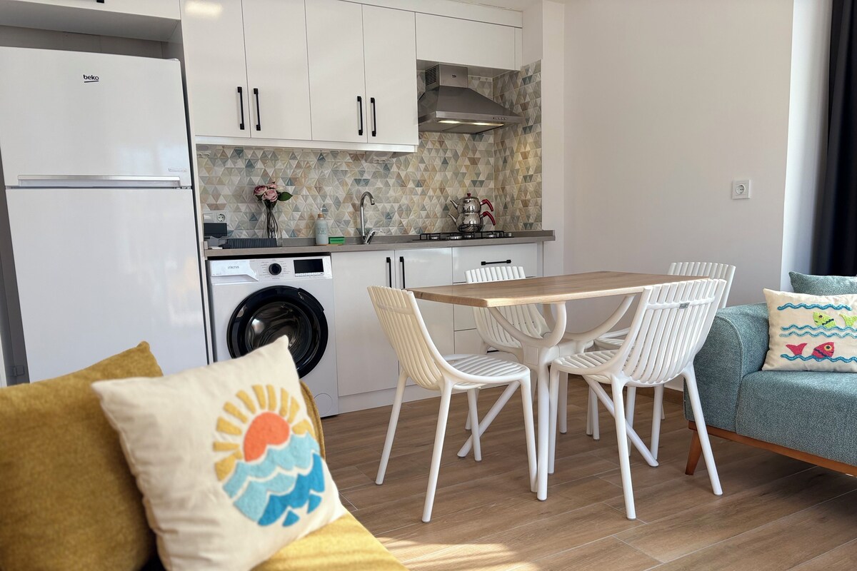 A modern kitchen with a built-in washing machine, a dining area with a wooden table and white chairs, and a sofa with a cushion.