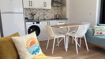 Una cocina moderna con lavadora integrada, un comedor con mesa de madera y sillas blancas, y un sofá con cojín.