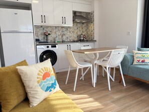 Una cocina moderna con lavadora integrada, un comedor con mesa de madera y sillas blancas, y un sofá con cojín.