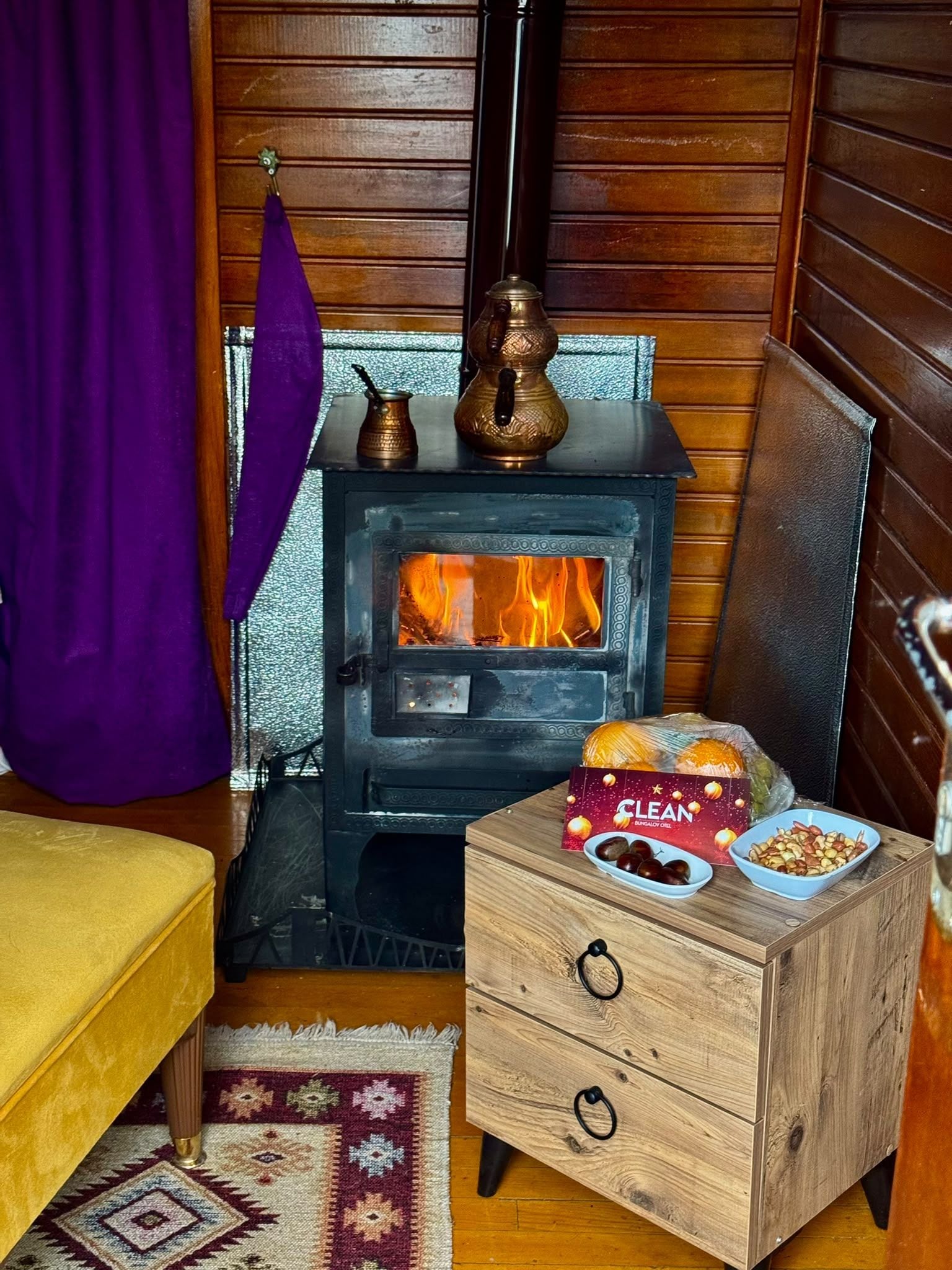 A cozy room with a wood-burning stove, a chest of drawers, and a small table with a box labeled 'CLEAN Bungalow Oil'.