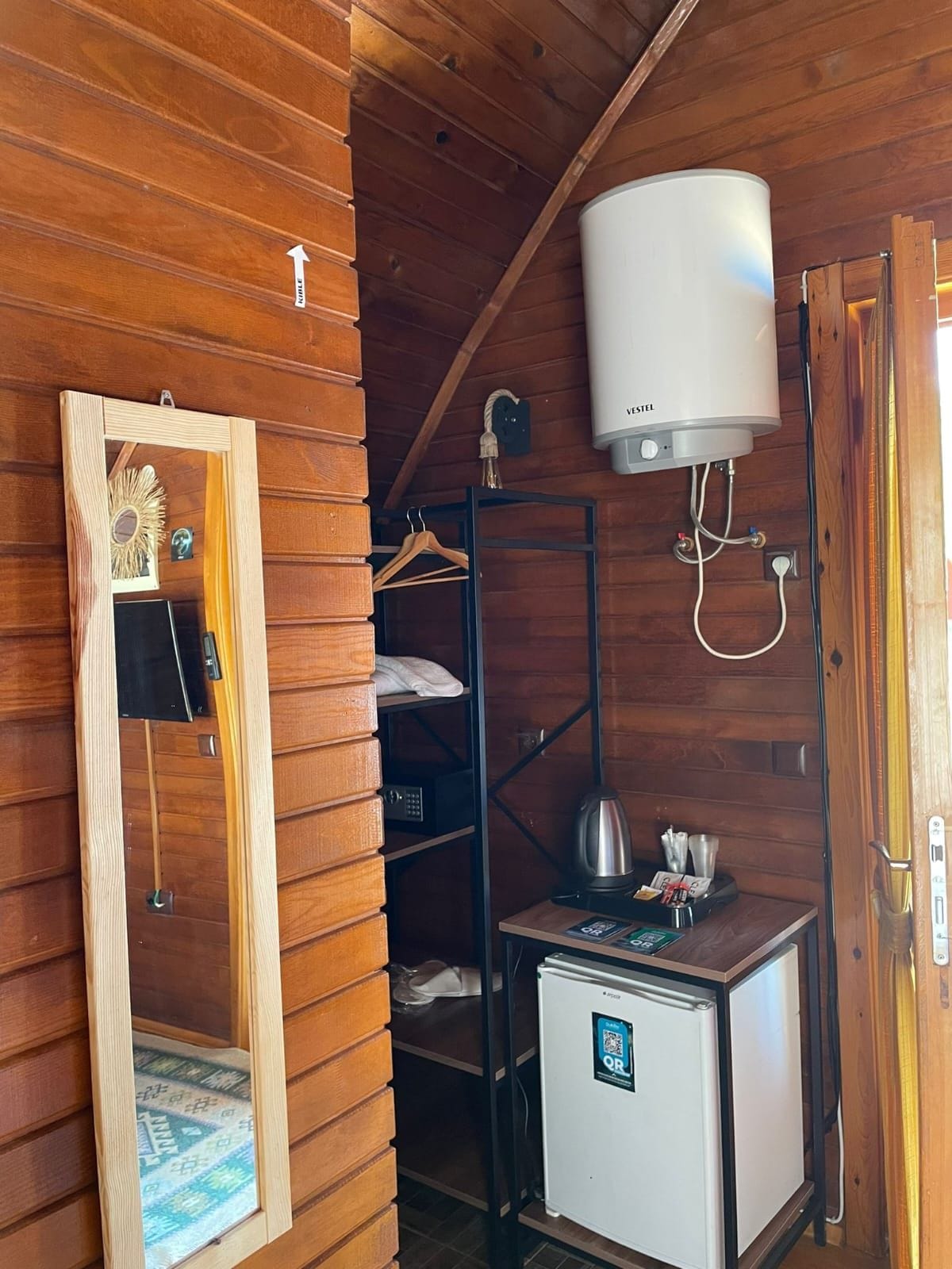 Quarto compacto com interior em madeira, aquecedor branco, pequena geladeira e espelho com moldura decorativa.