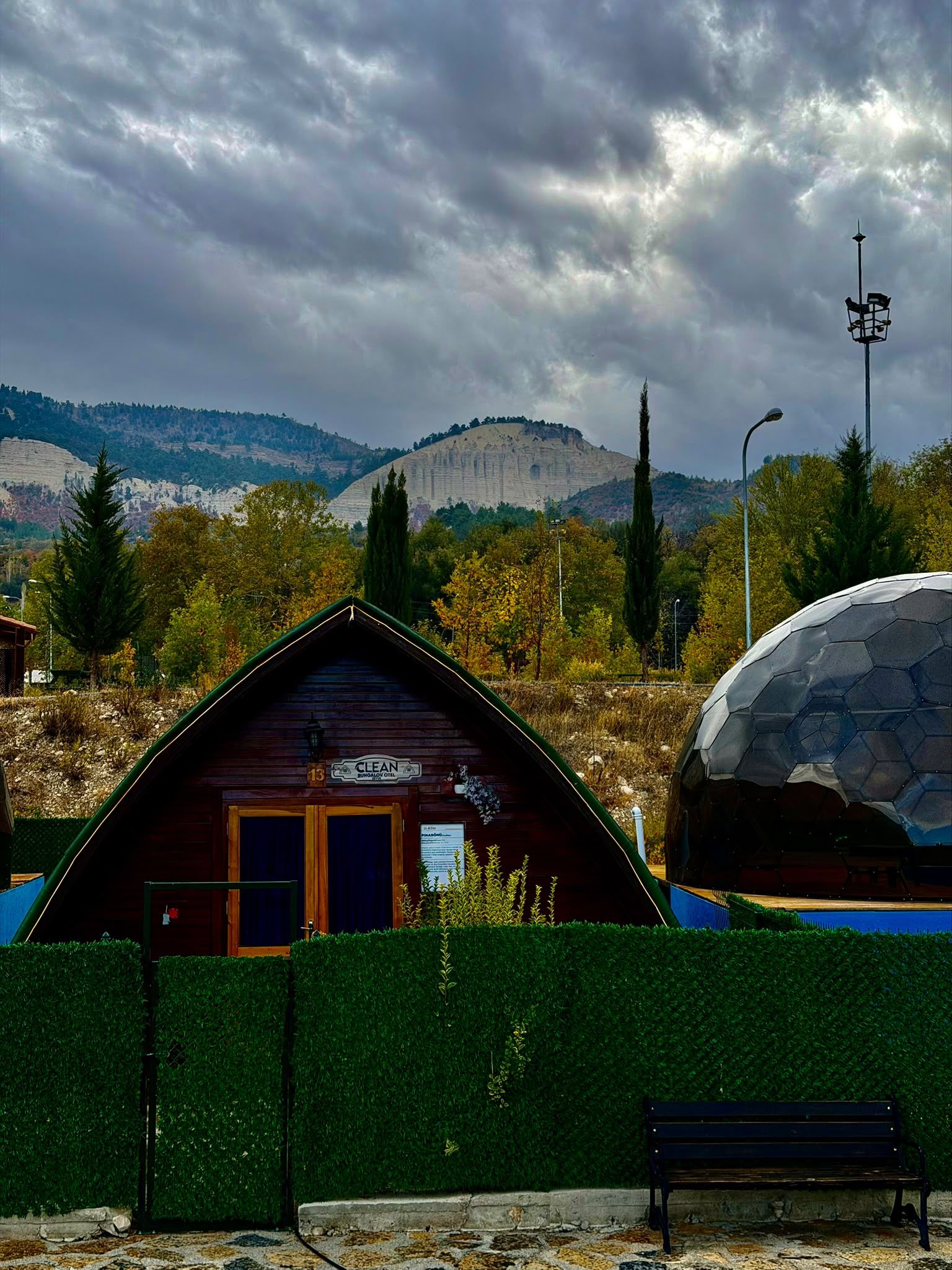 A wooden cabin with a sign reading 'CLEAN BUNGALOW COTTLE,' surrounded by a green hedge, with a bench and a large dome structure in the background.