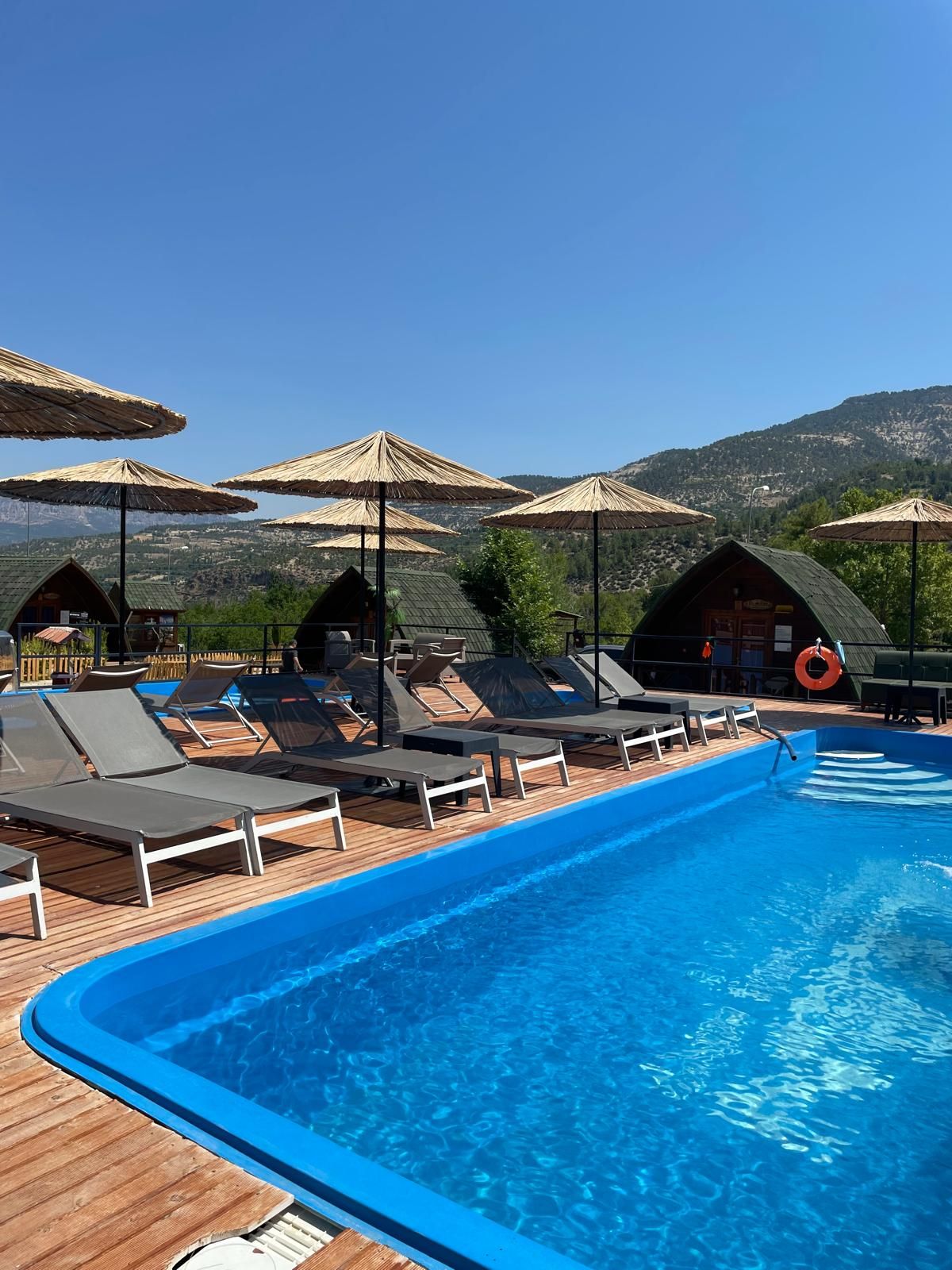 A swimming pool with lounge chairs and umbrellas, surrounded by wooden deck and hills in the background.