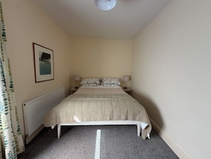 Ein Schlafzimmer mit einem Bett, zwei Nachttischlampen, einer Heizung, einem gerahmten Bild an der Wand und einem Vorhang mit Blätermotiv.