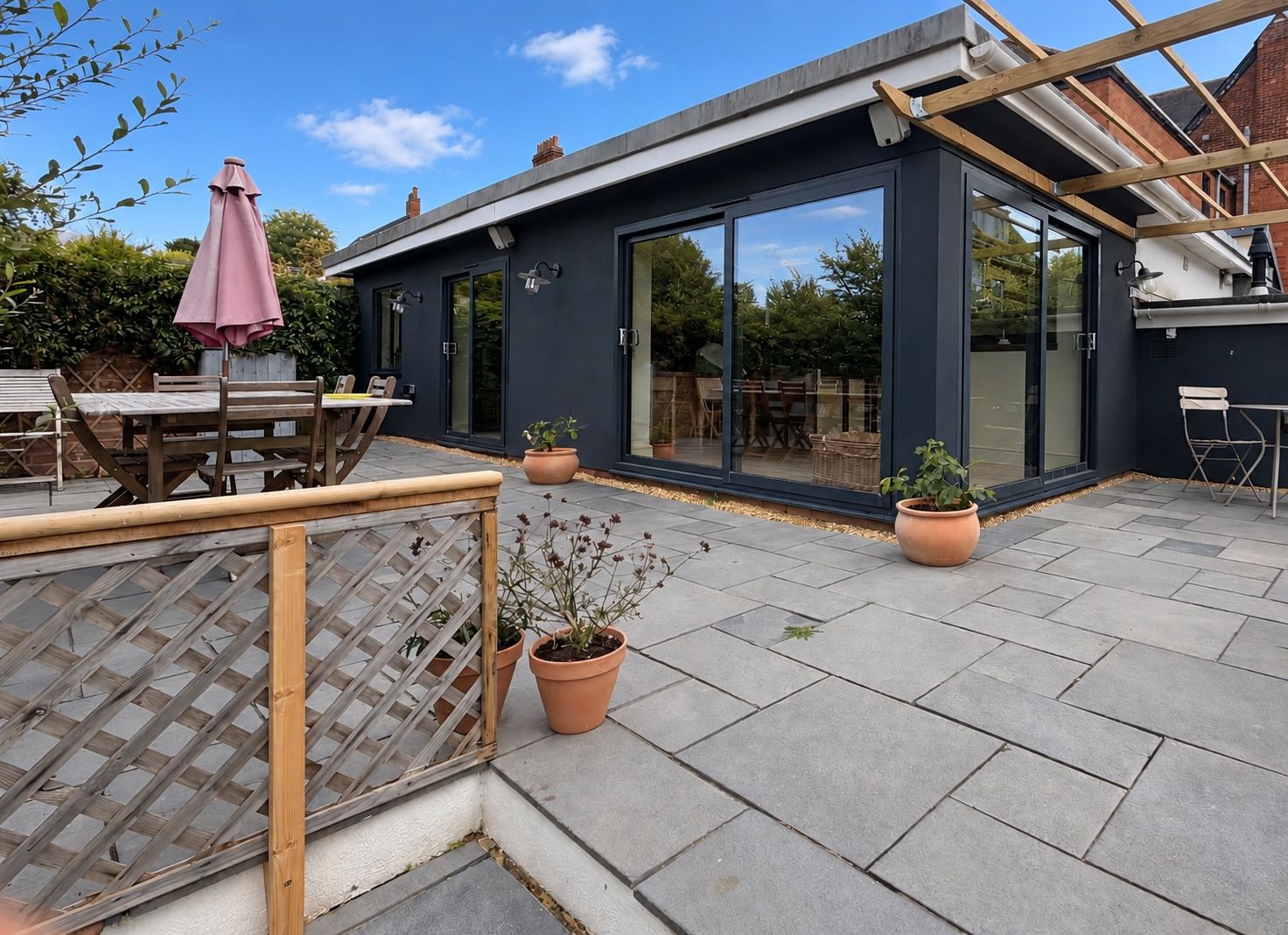A modern patio with a wooden table and chairs, potted plants, and a glass door leading to an outdoor dining area.