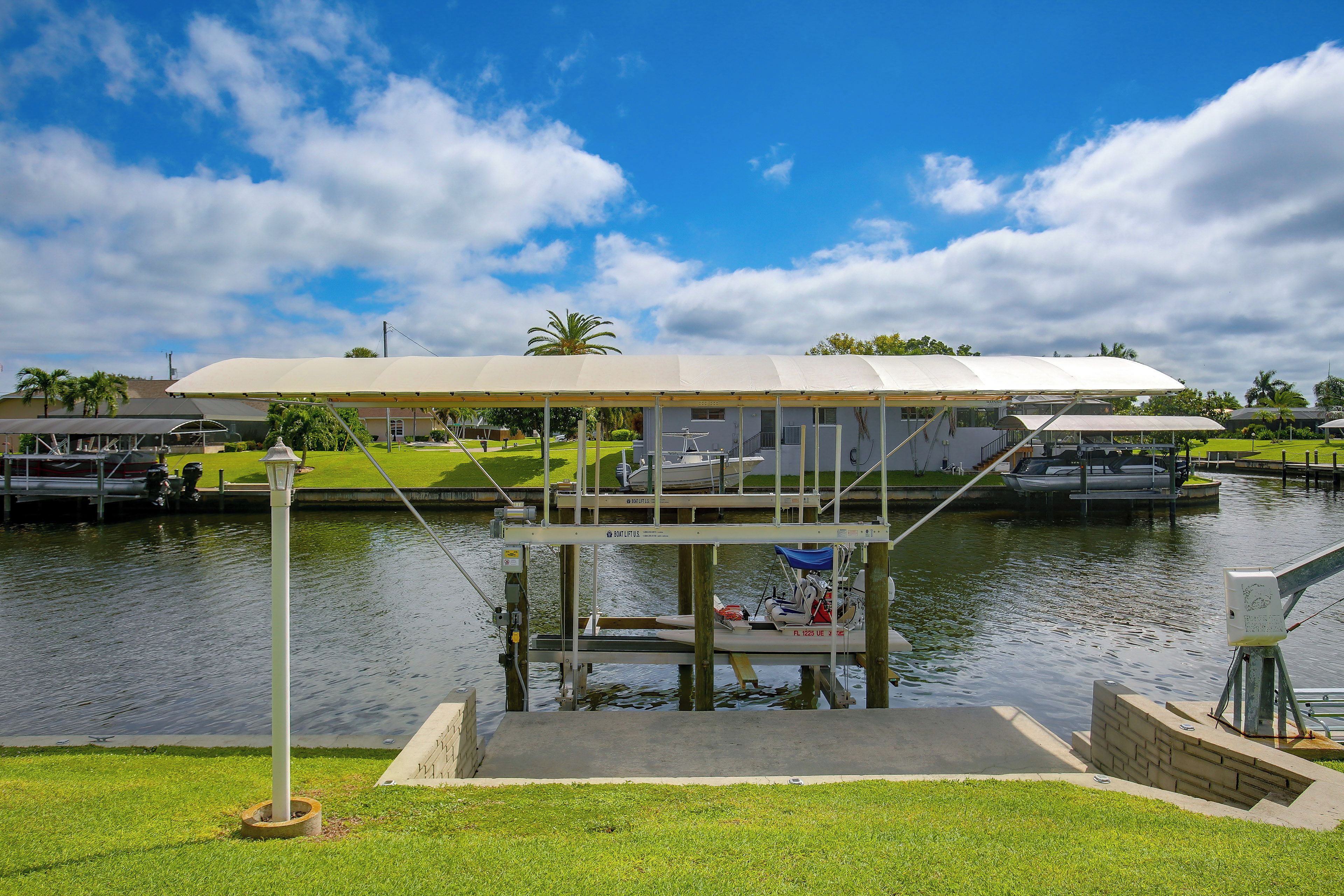 A covered boat lift structure with a grassy area in the foreground and a body of water with boats in the background.