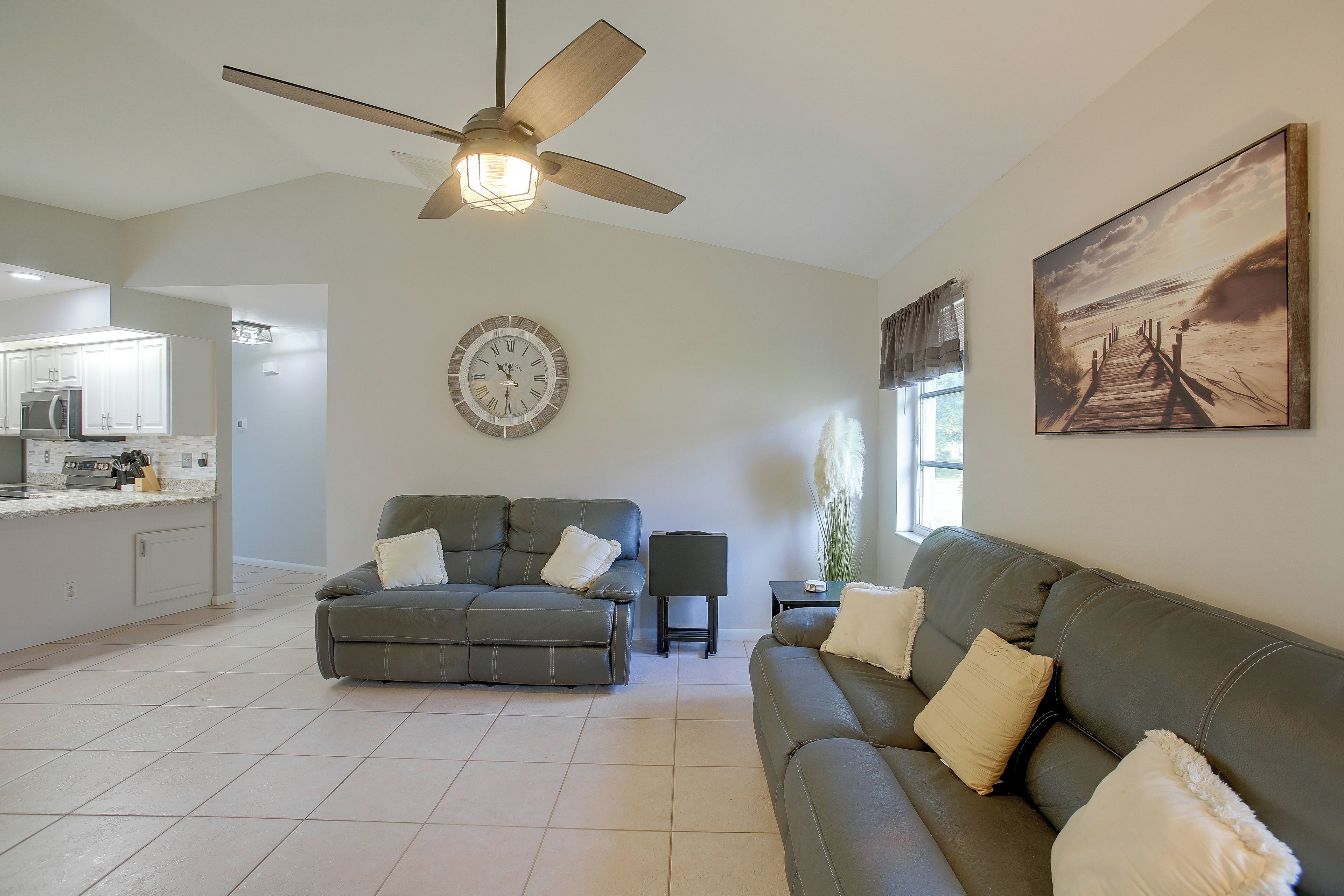A living room with a ceiling fan, a clock, a couch, a dining area with a microwave, and a painting on the wall.
