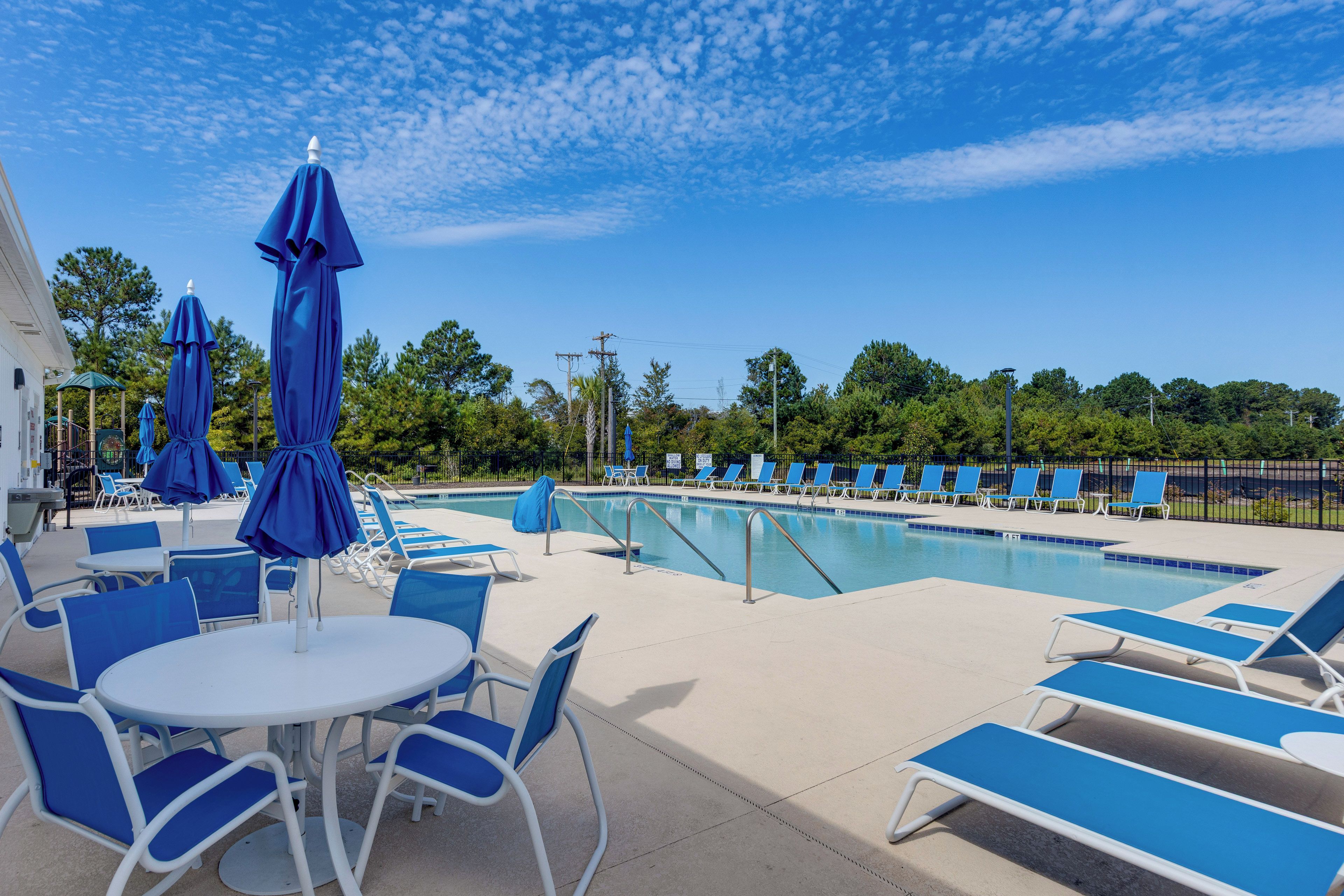 A poolside area with lounge chairs and umbrellas, a pool, and a view of trees and a building in the background.