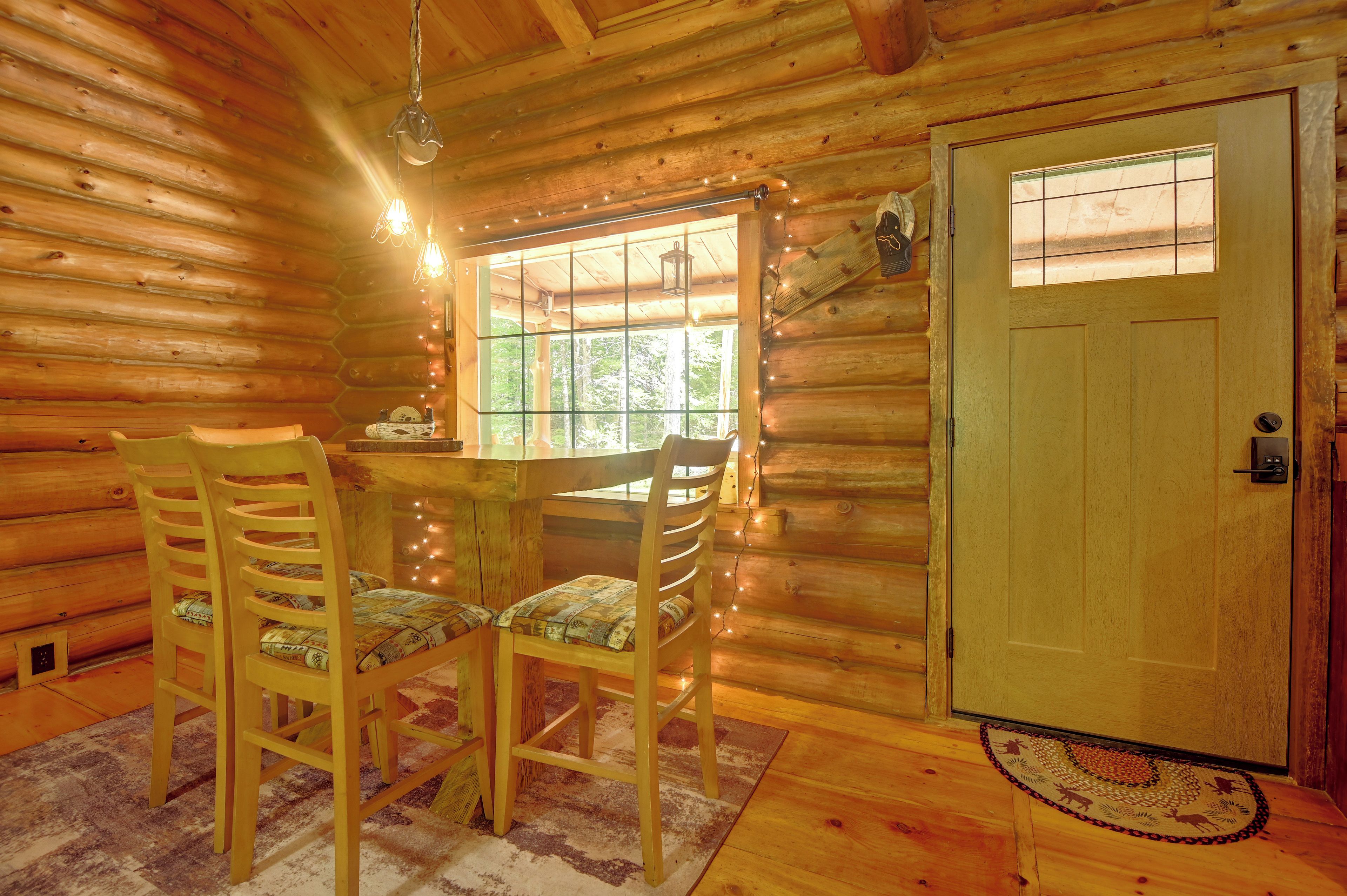 Interior de una cabaña de madera con una mesa de comedor, dos sillas, una ventana con cortinas, una puerta y un lámpara colgante.