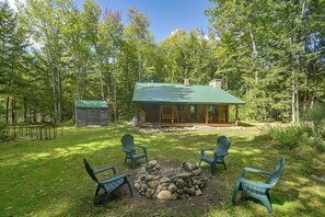 A cabin with a green roof surrounded by a forest, a fire pit with rocks, and four Adirondack chairs. - Maine Log Cabin! Private 2.5-acre Getaway! (Denmark)