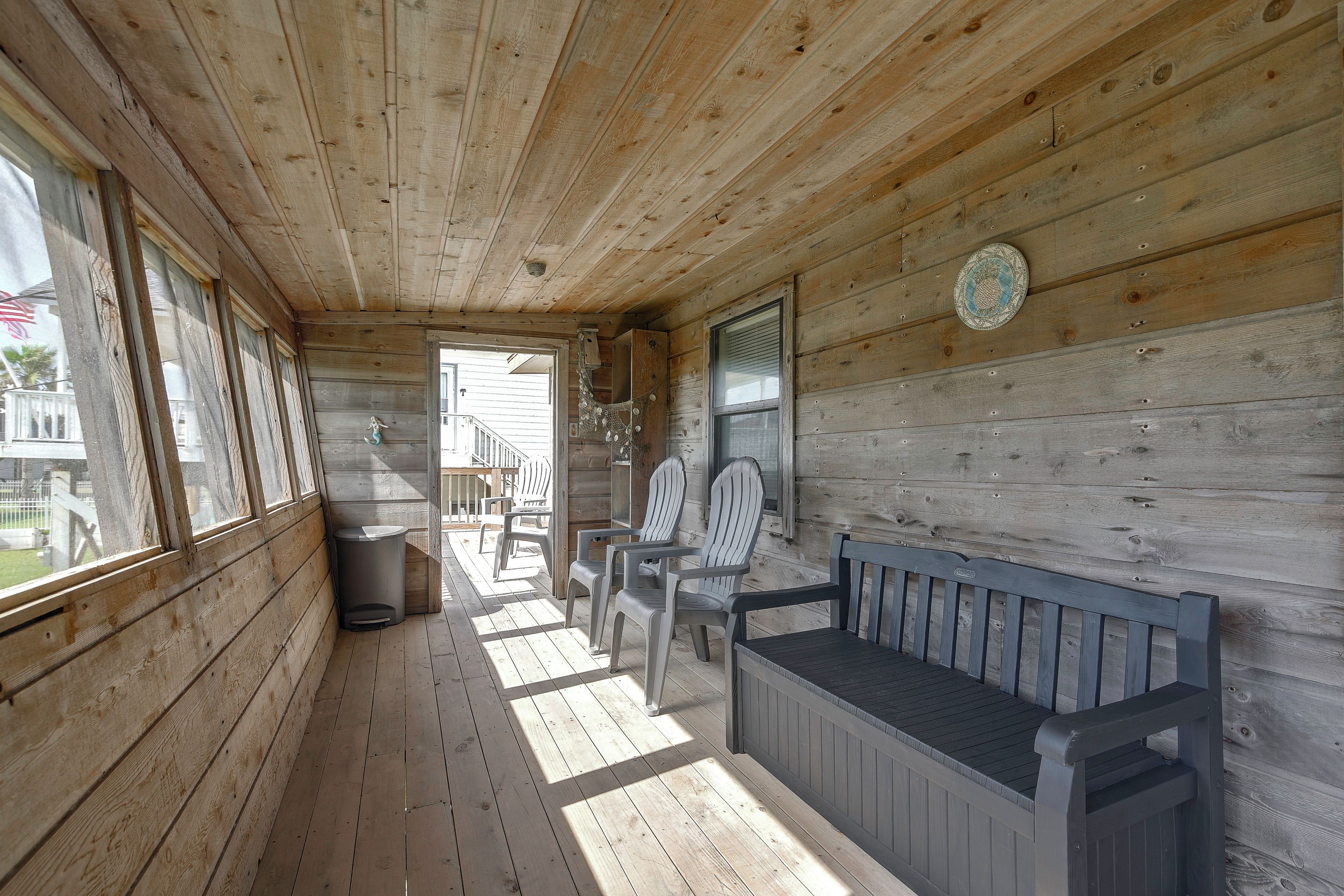 A wooden porch with a bench and chairs, a trash can, and a view of the outside.