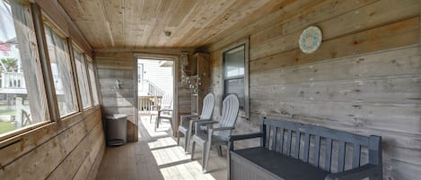 A wooden porch with a bench and chairs, a trash can, and a view of the outside.