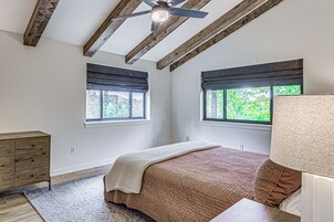 A bedroom with a wooden ceiling, two windows with blinds, a bed with a patterned bedspread, a bedside table, and a dresser.