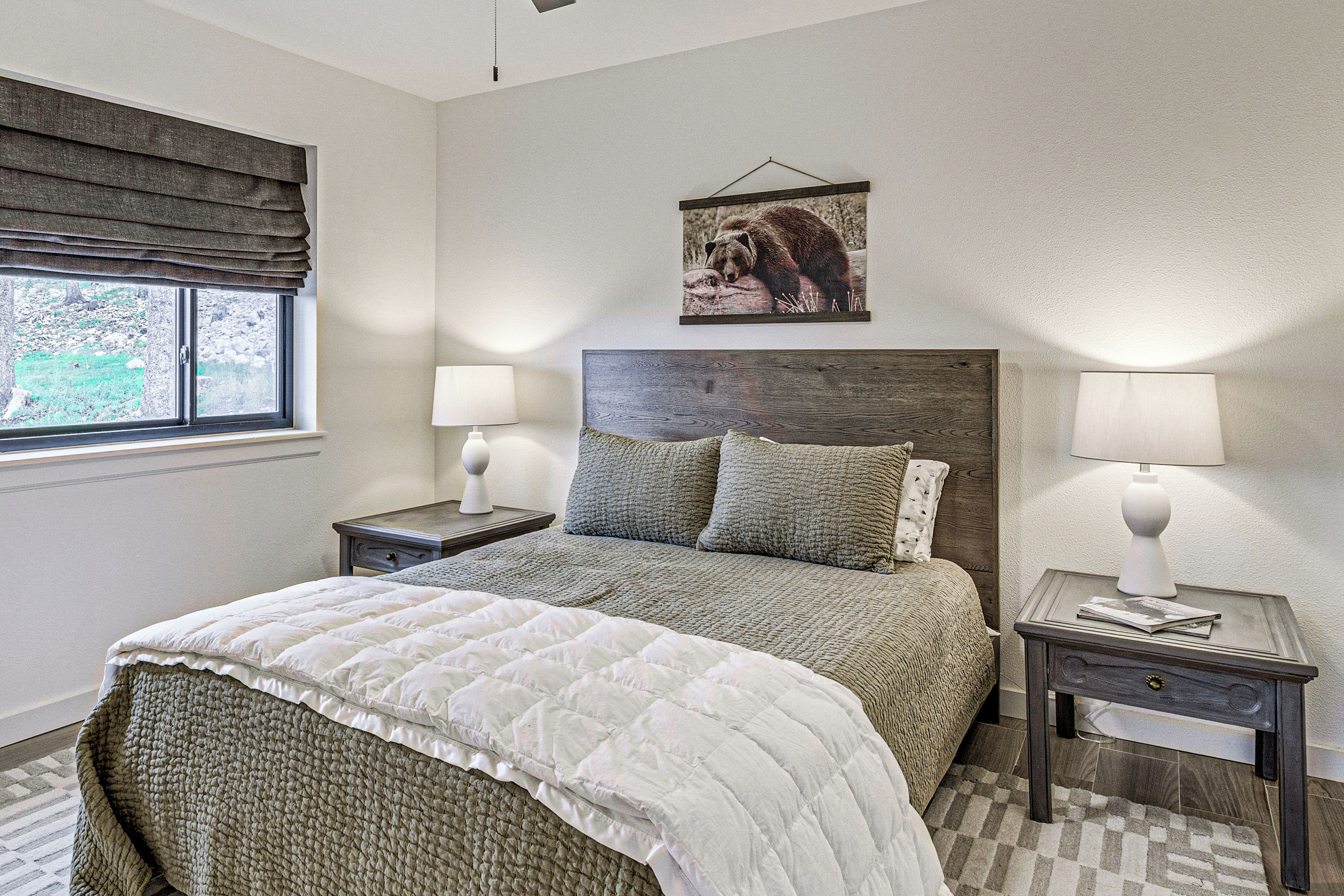 A bedroom with a bed, two bedside tables, a window with blinds, a wall-mounted picture, and a textured rug.
