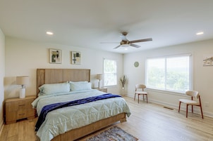 Quarto com cama, mesas de cabeceira, ventilador de teto e janelas com persianas.