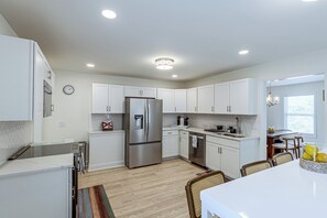 A modern kitchen with white cabinetry, stainless steel appliances, and a dining area with a table and chairs. - Deck & Game Room! Modern Lake Guntersville Retreat (Guntersville)