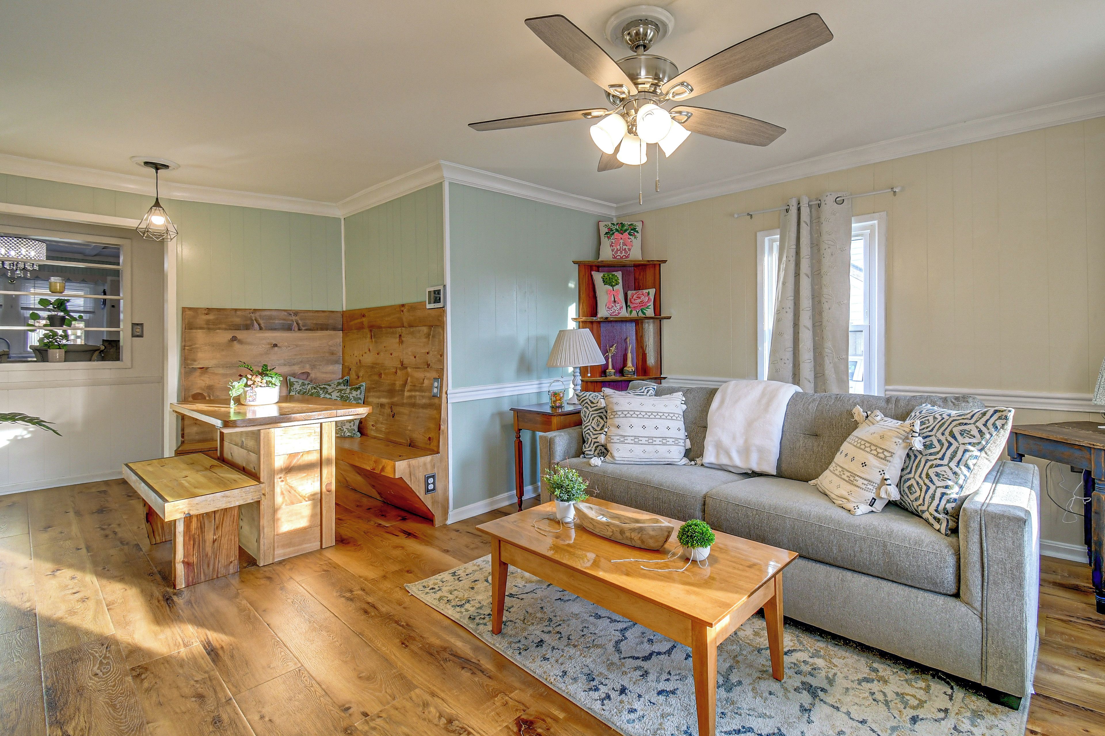 A cozy living room with a wooden coffee table, a sofa with patterned cushions, a ceiling fan, and a window with curtains.