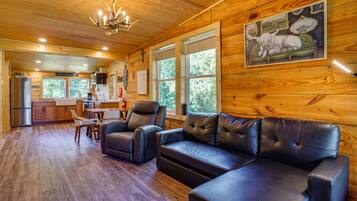 A cabin interior with a wooden ceiling, a dining area, a kitchen, and a living room with leather sofas and a painting of a white animal.