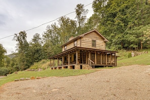 A two-story log cabin with a porch, surrounded by a forested hillside.