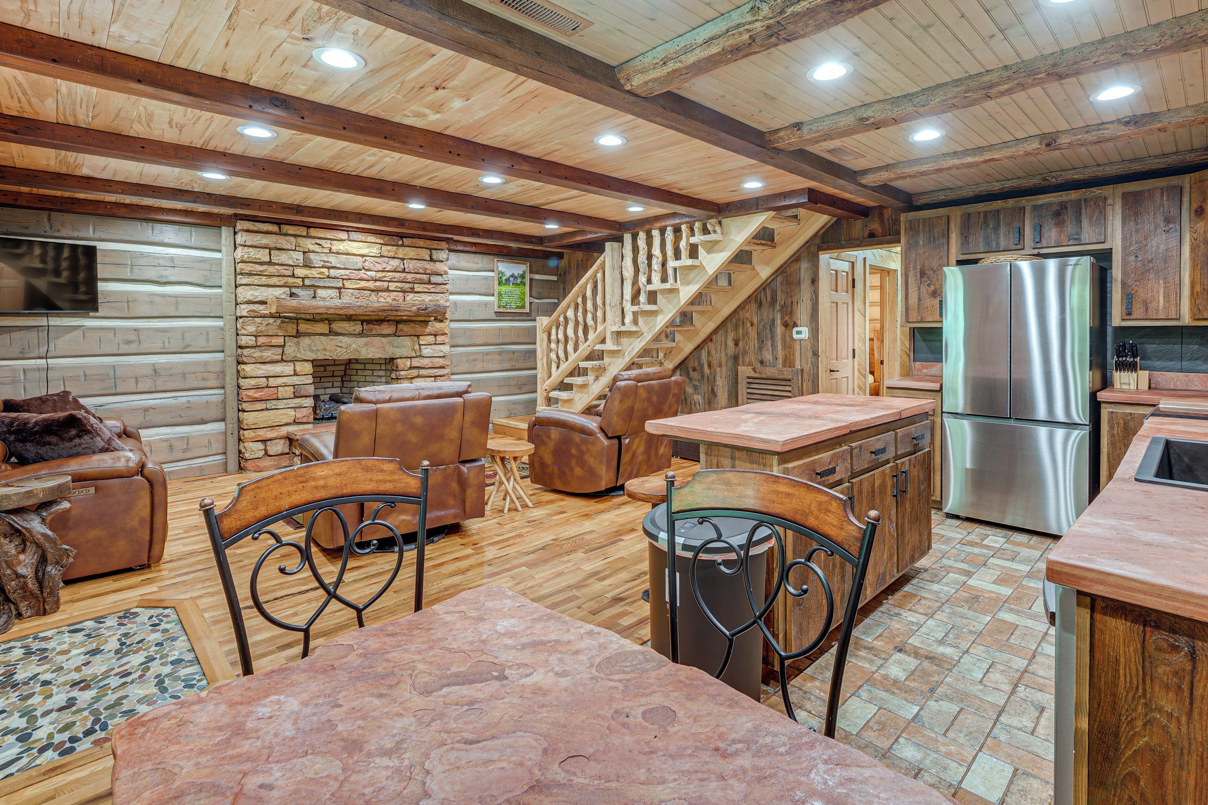 A rustic interior with a stone fireplace, wooden staircase, leather seating, and a stainless steel refrigerator.