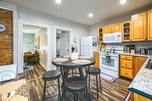A modern kitchen with wooden cabinets, a white refrigerator, a microwave, and a stove. There is a dining area with a round table and stools. - Modern 1br: Queen Bed, Coffee Bar & Walkin Shower (Parkersburg)