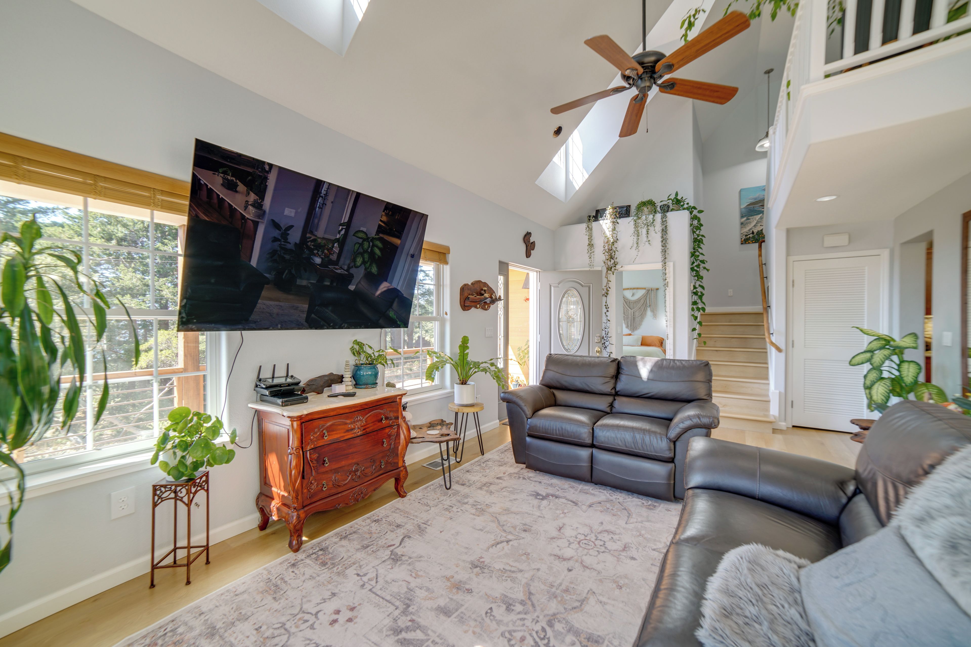 A spacious living room with a ceiling fan, a large flat-screen TV, leather sofas, and a variety of potted plants.