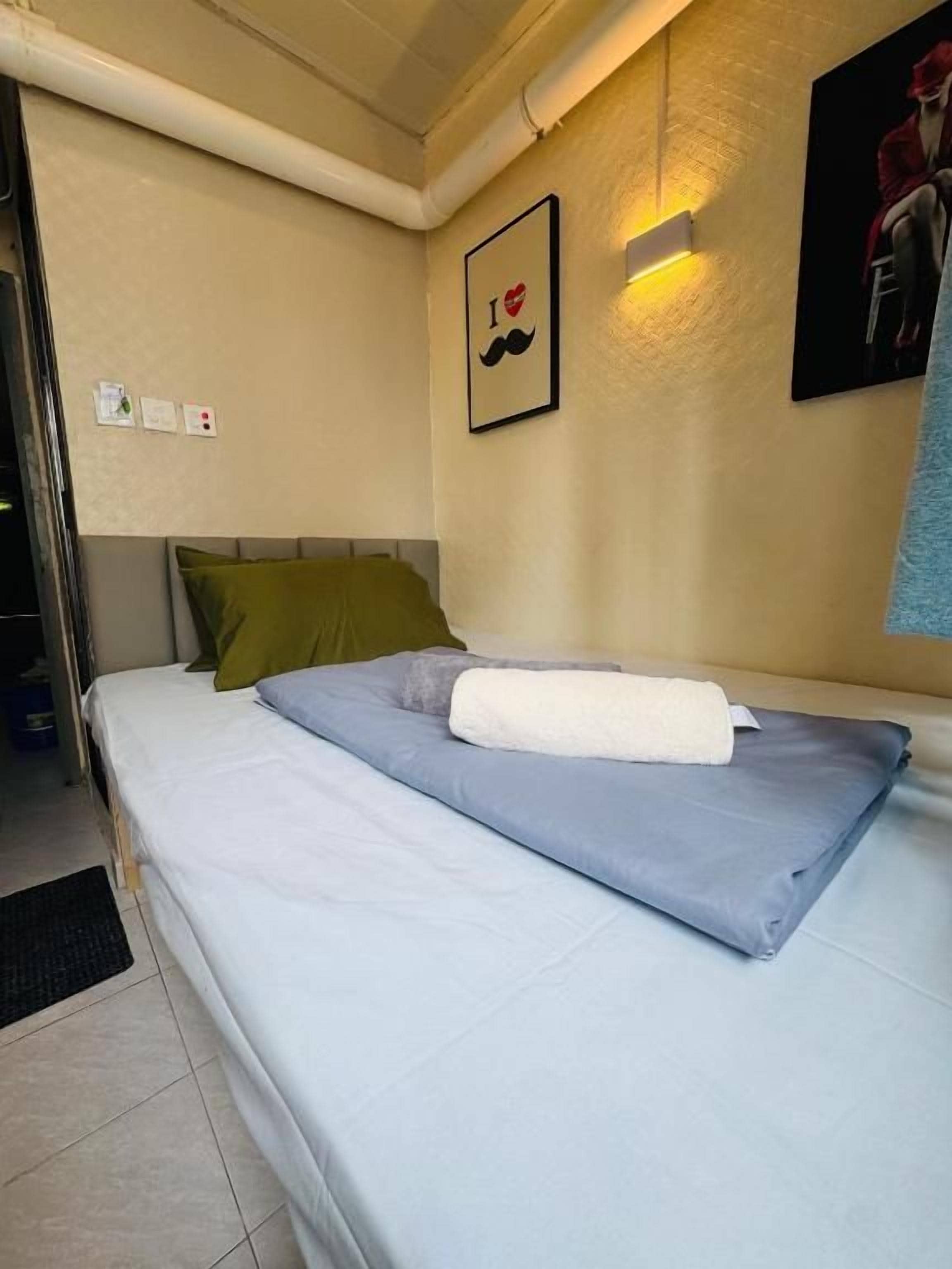 Un lit simple avec un matelas gris, une couverture blanche et un oreiller vert. Un tableau encadré représentant un cœur et une moustache est accroché au mur.
