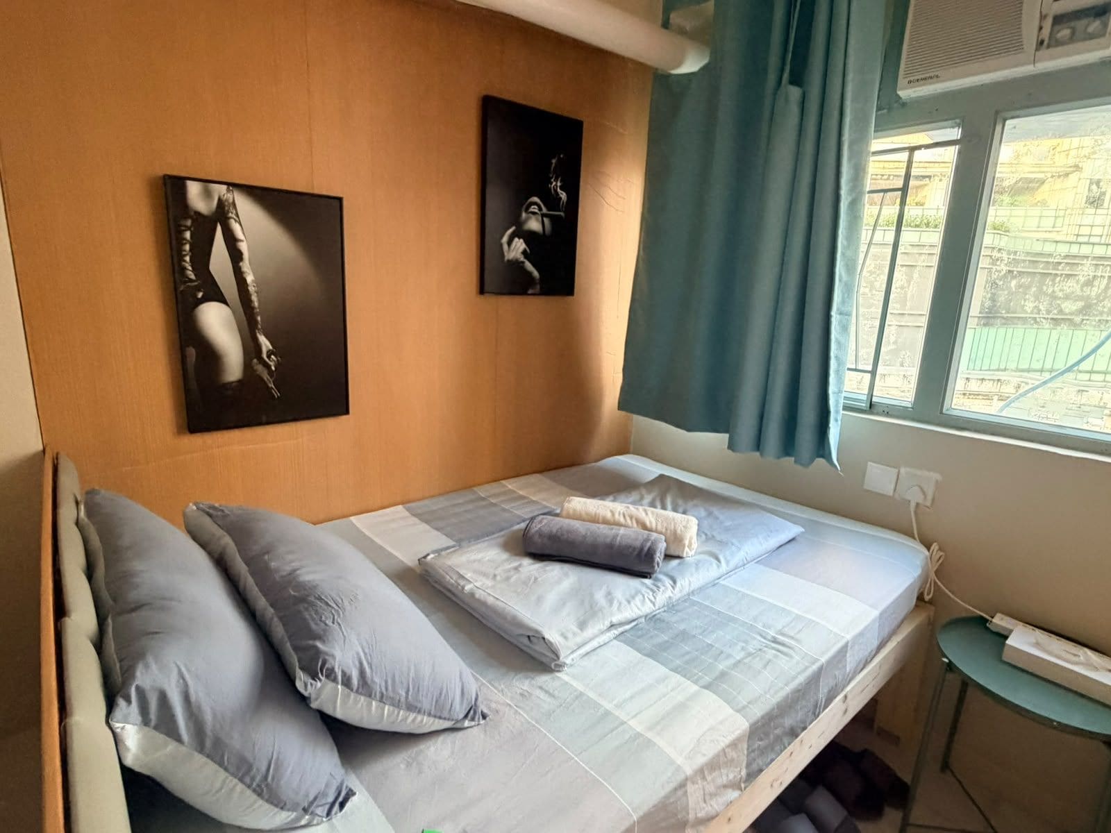 A single bed with a striped bedspread and pillows, a wall-mounted air conditioner, and a window with curtains.