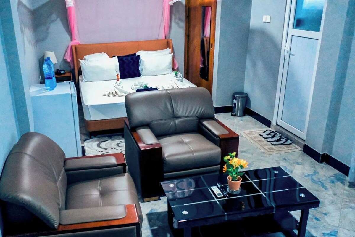 Ein Hotelzimmer mit einem Bett, zwei Ledersesseln, einem kleinen Tisch mit einer Vase und einem Nachttisch mit einer Lampe.