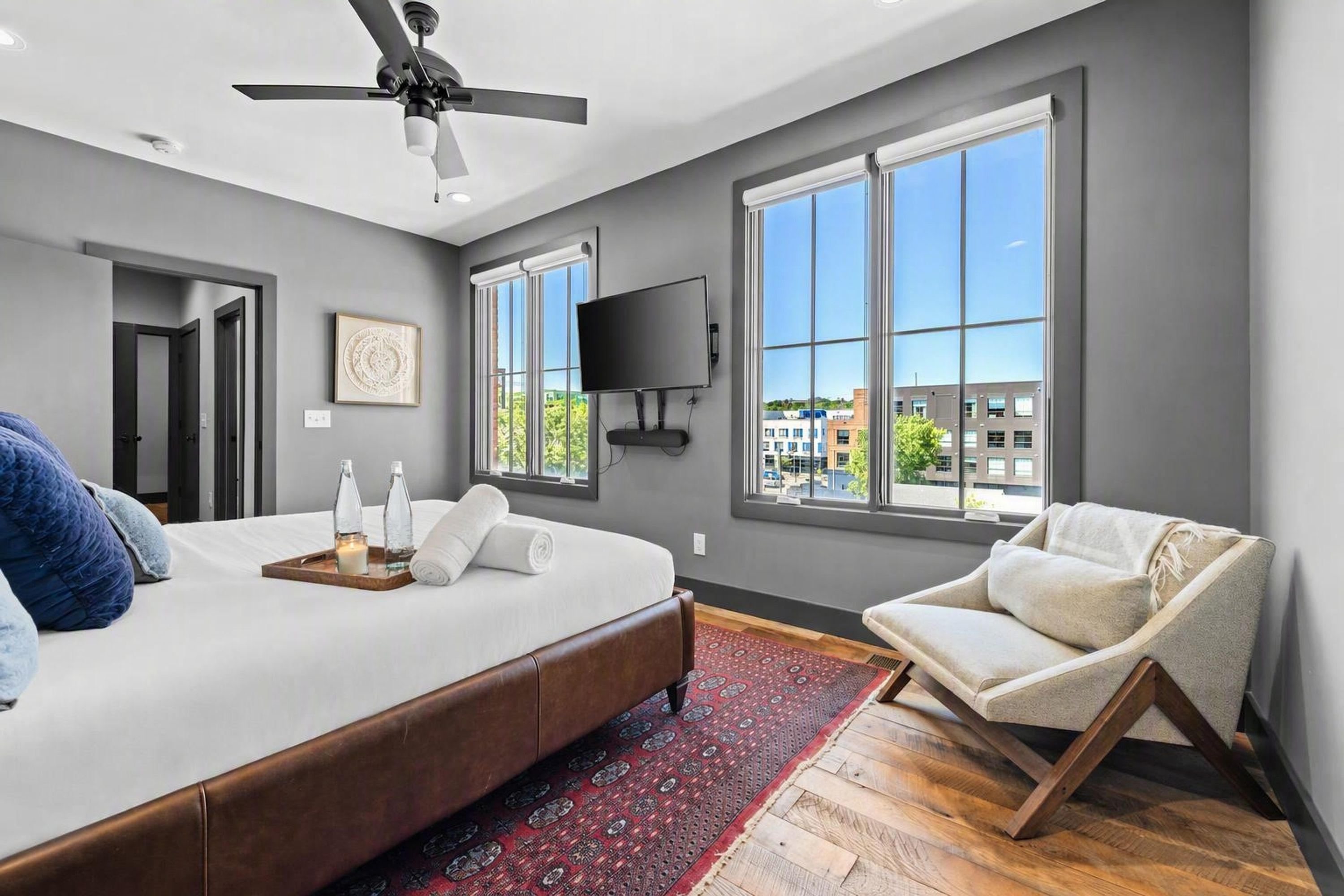 A modern bedroom with a large bed, a ceiling fan, a TV, and a view of buildings through large windows.