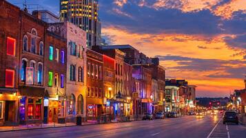 A street lined with historic buildings at dusk, featuring illuminated storefronts and a modern skyscraper in the background.