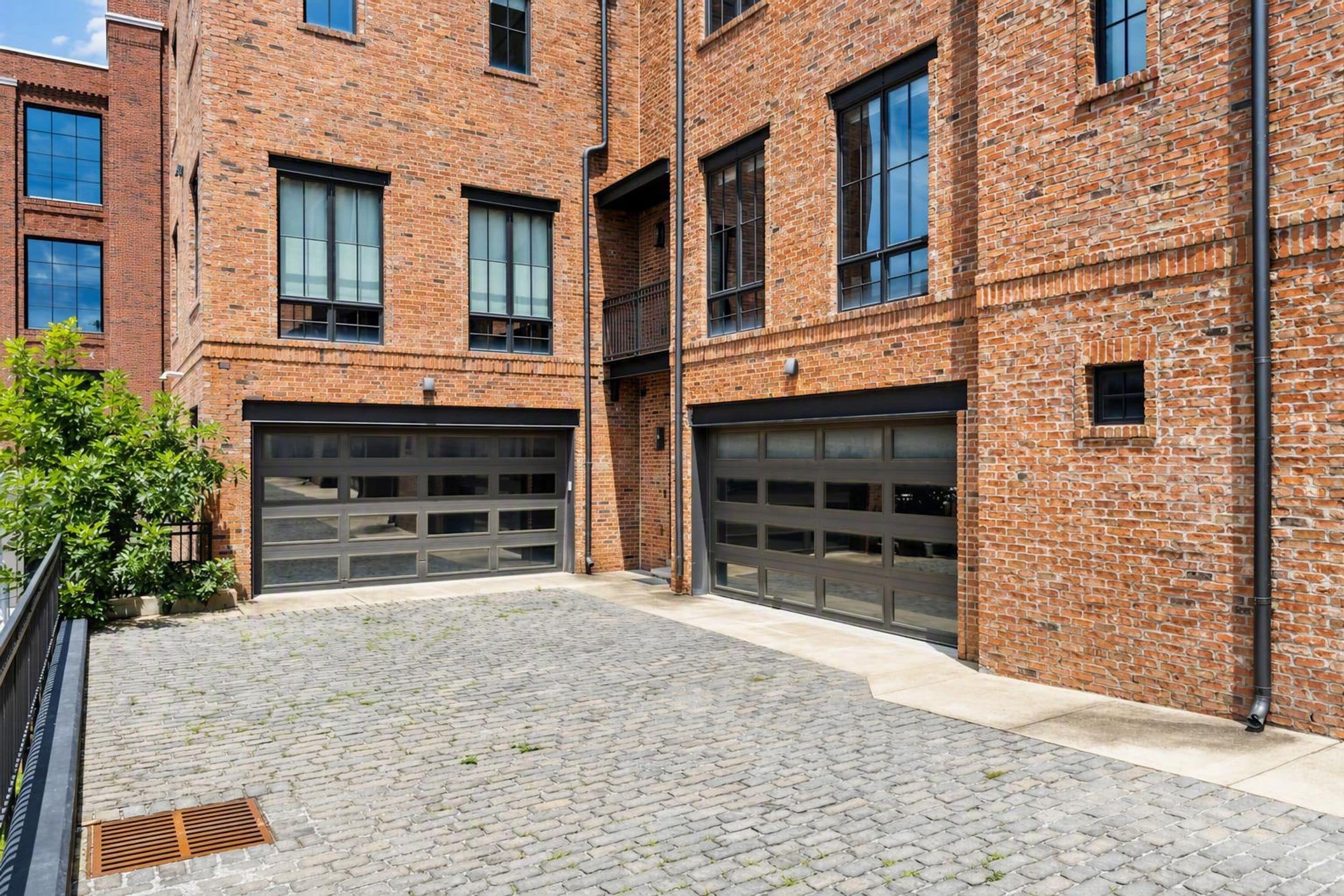 A brick building with two garage doors, a paved courtyard, and a metal fence.