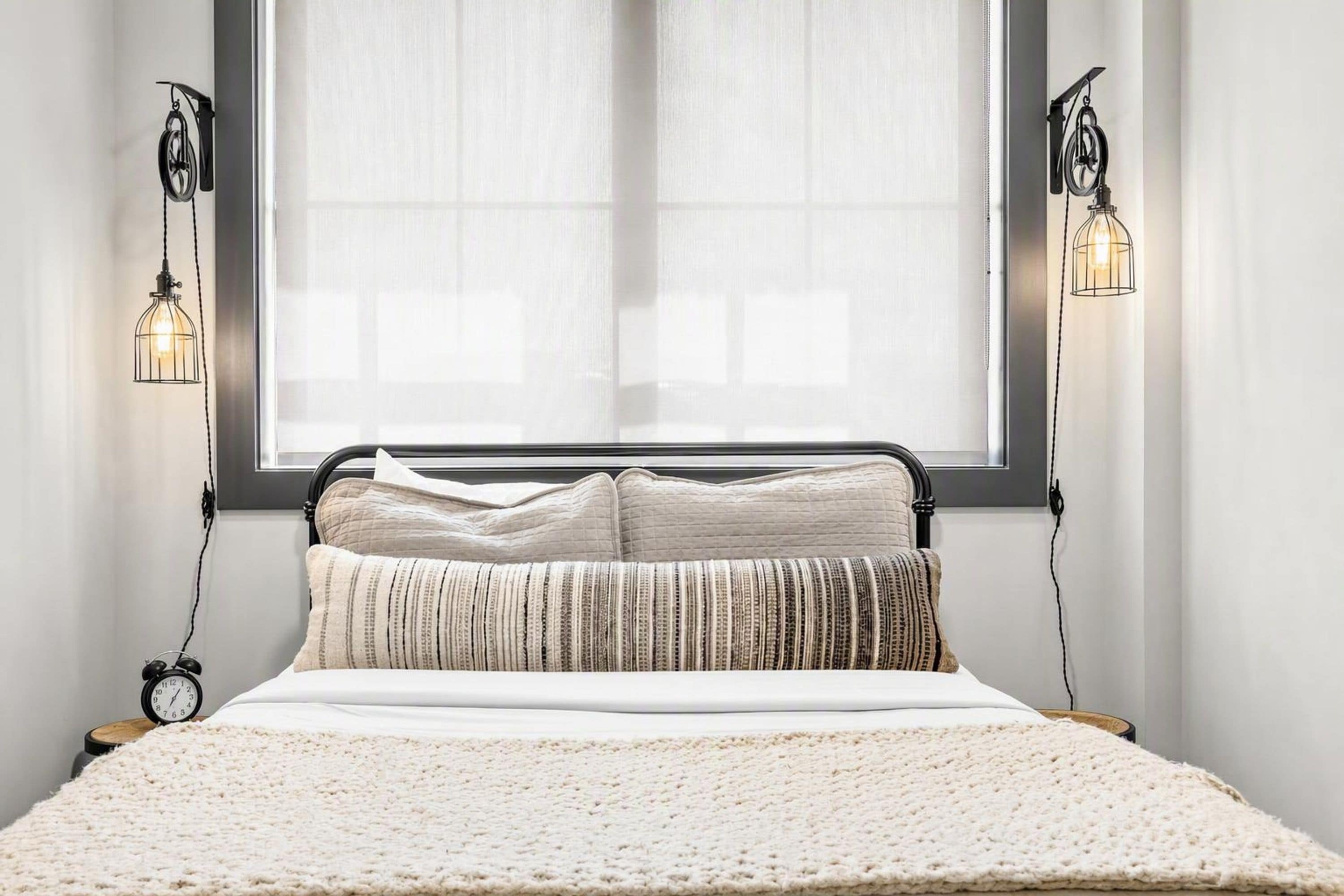 Een bed met een witte deken en twee kussens, een zwart metalen hoofdbord, een raam met dunne gordijnen en twee wandlampen.
