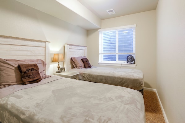 A bedroom with two beds, a nightstand with a lamp, a window with blinds, and a ceiling fan.