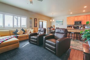 A living room with a sofa, two leather chairs, a coffee table, and a dining area with a table and chairs. There is a kitchen area with cabinets and appliances.
