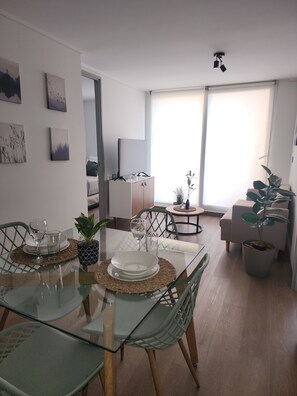 Ein modernes Wohnzimmer mit einem Glastisch, Stühlen und einer Couch. An den Wänden befinden sich Gemälde und eine Deckenleuchte.