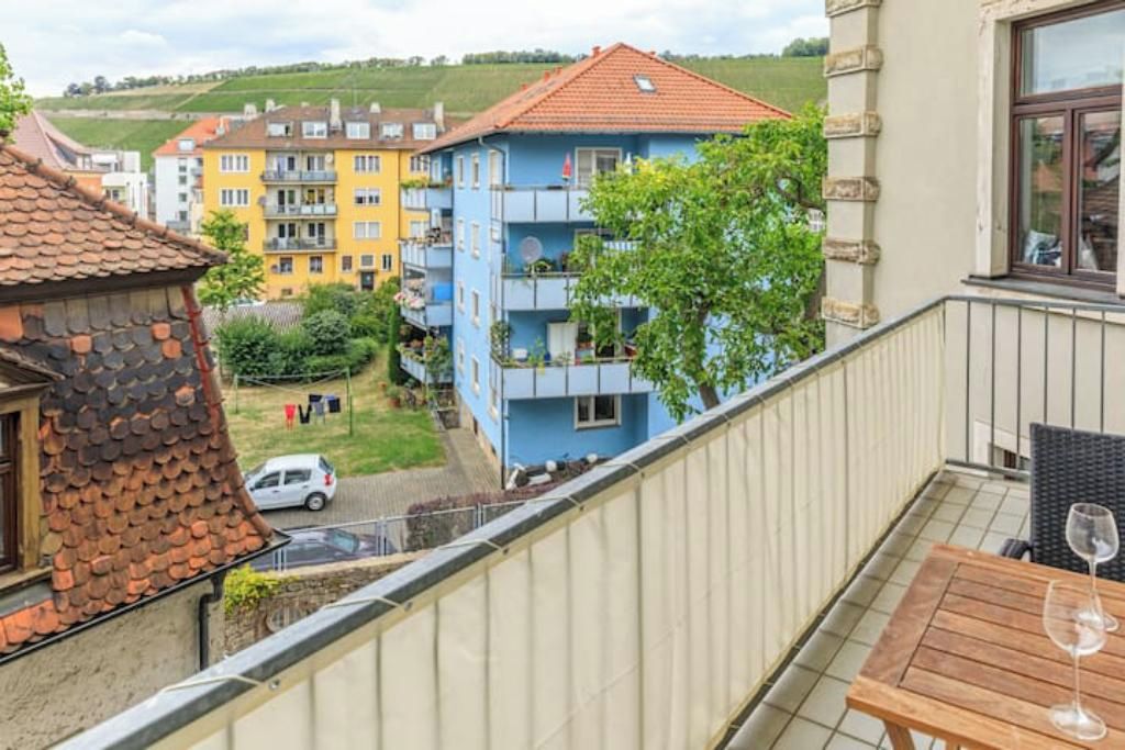 Ein Balkon mit Tisch und Weinglas, mit Blick auf ein Wohngebiet mit farbenfrohen Gebäuden und einem geparkten Auto auf der Straße.