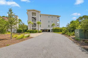 A multi-story building with a central entrance, a parking area, and surrounding greenery. - Moorings 101 – Marshview Sanctuary | 3BR/3BA • Pool • Screened Porch (Jekyll Island)