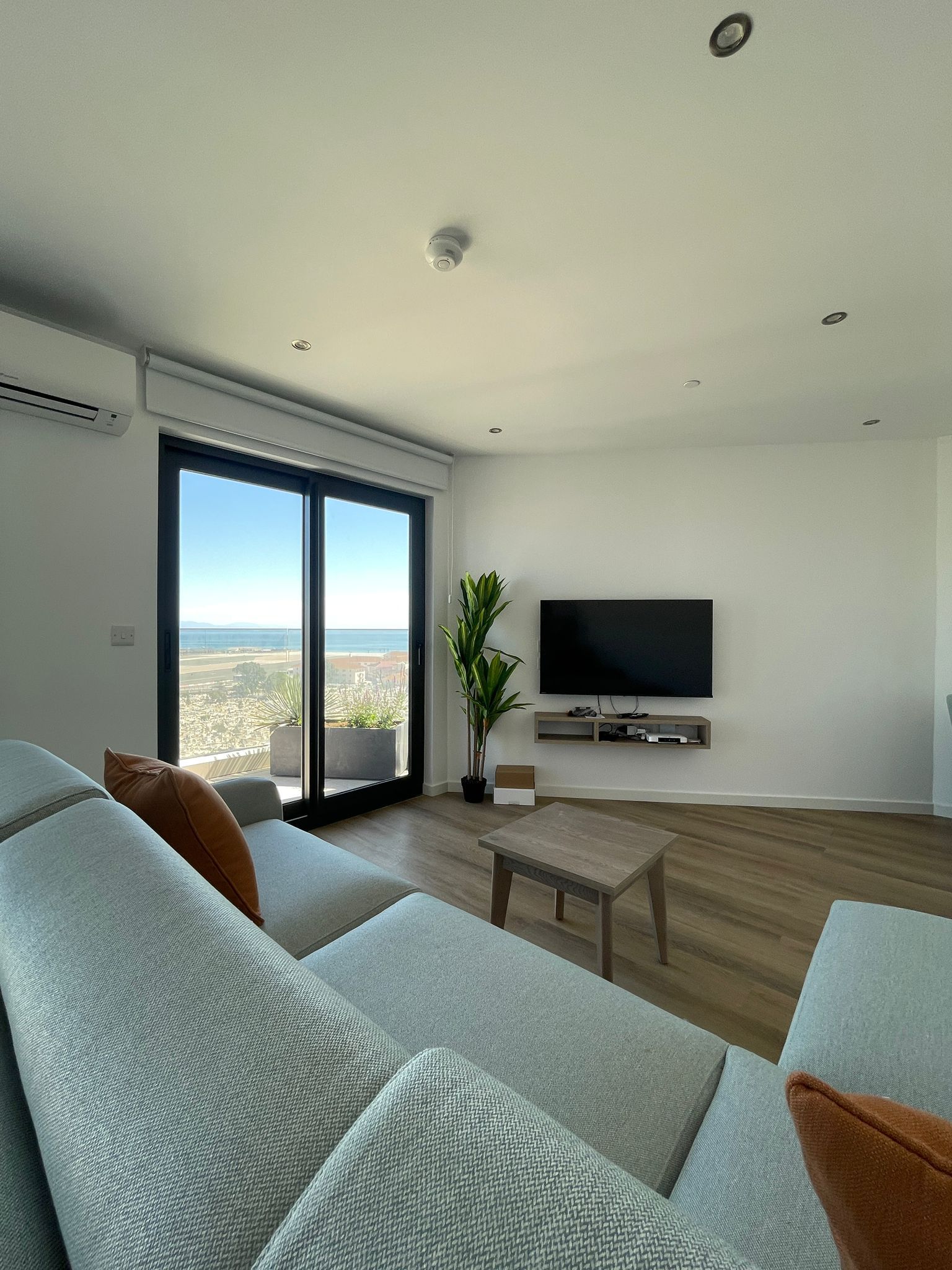 A modern living room with a large flat-screen TV, a sofa set, a coffee table, and a view of the city through floor-to-ceiling windows.