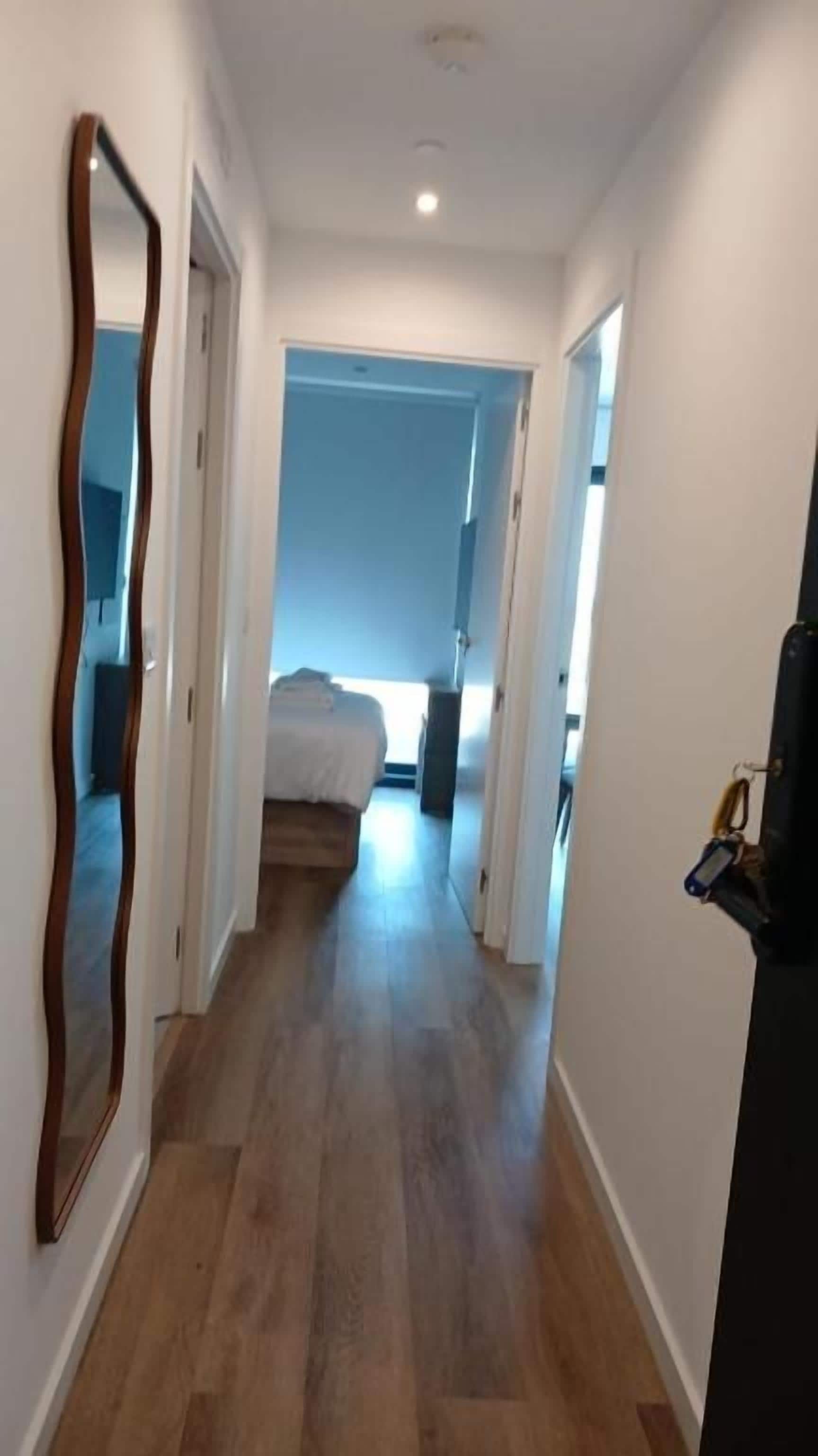 A narrow hallway with wooden flooring, leading to a bedroom with a bed and a window.